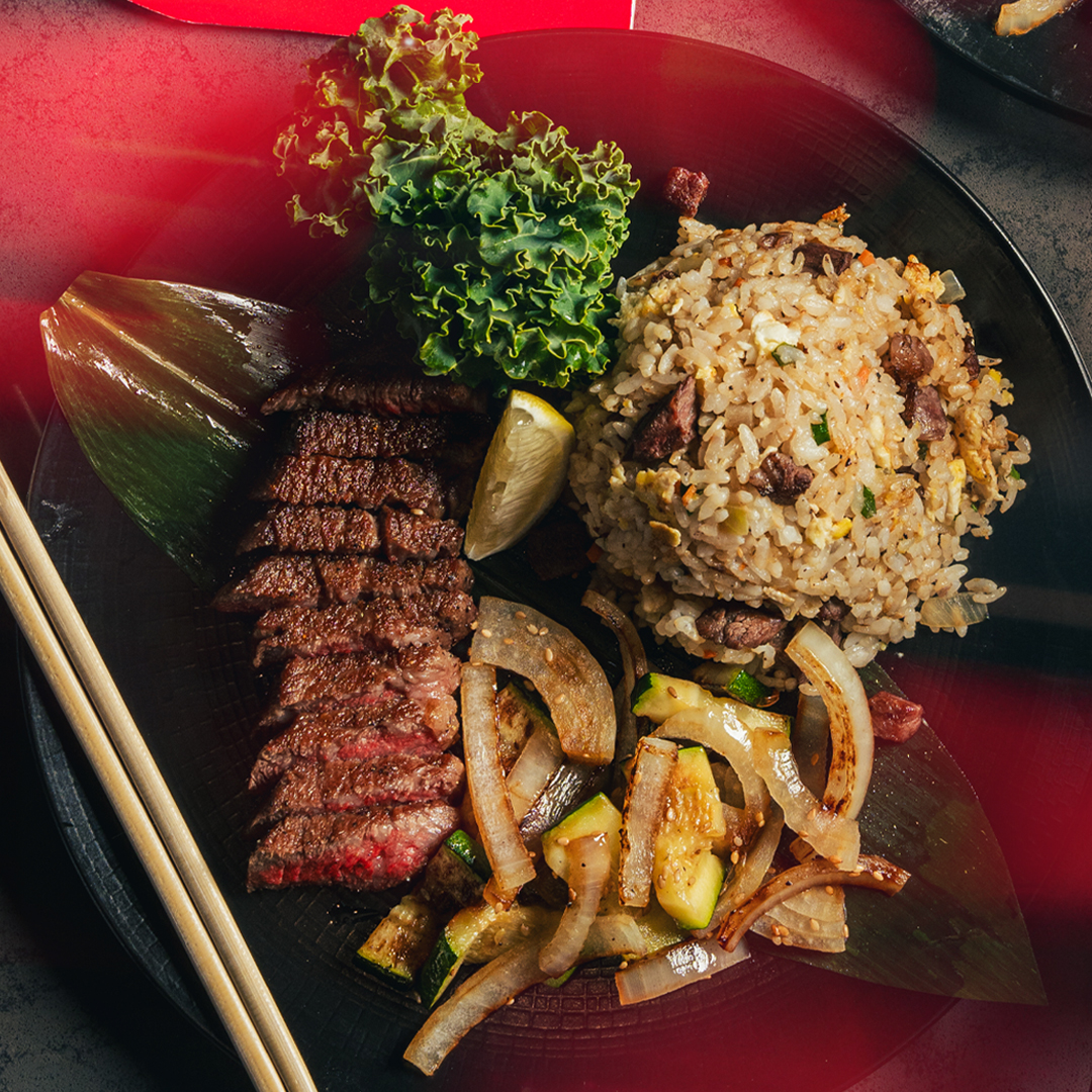 Sliced steak, fried rice, sautéed onions and zucchini, and greens arranged on a black plate.