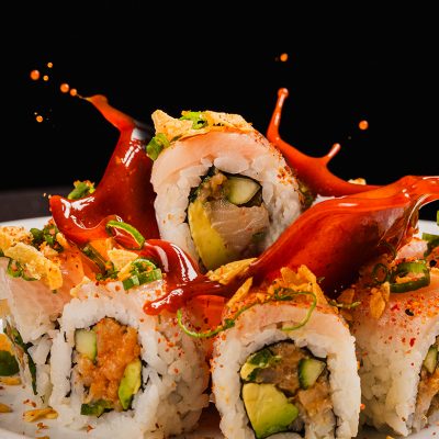 Sushi rolls filled with avocado and vegetables are topped with crispy flakes, sliced green onions. One landing with some vibrant red sauce., making a spalash.
