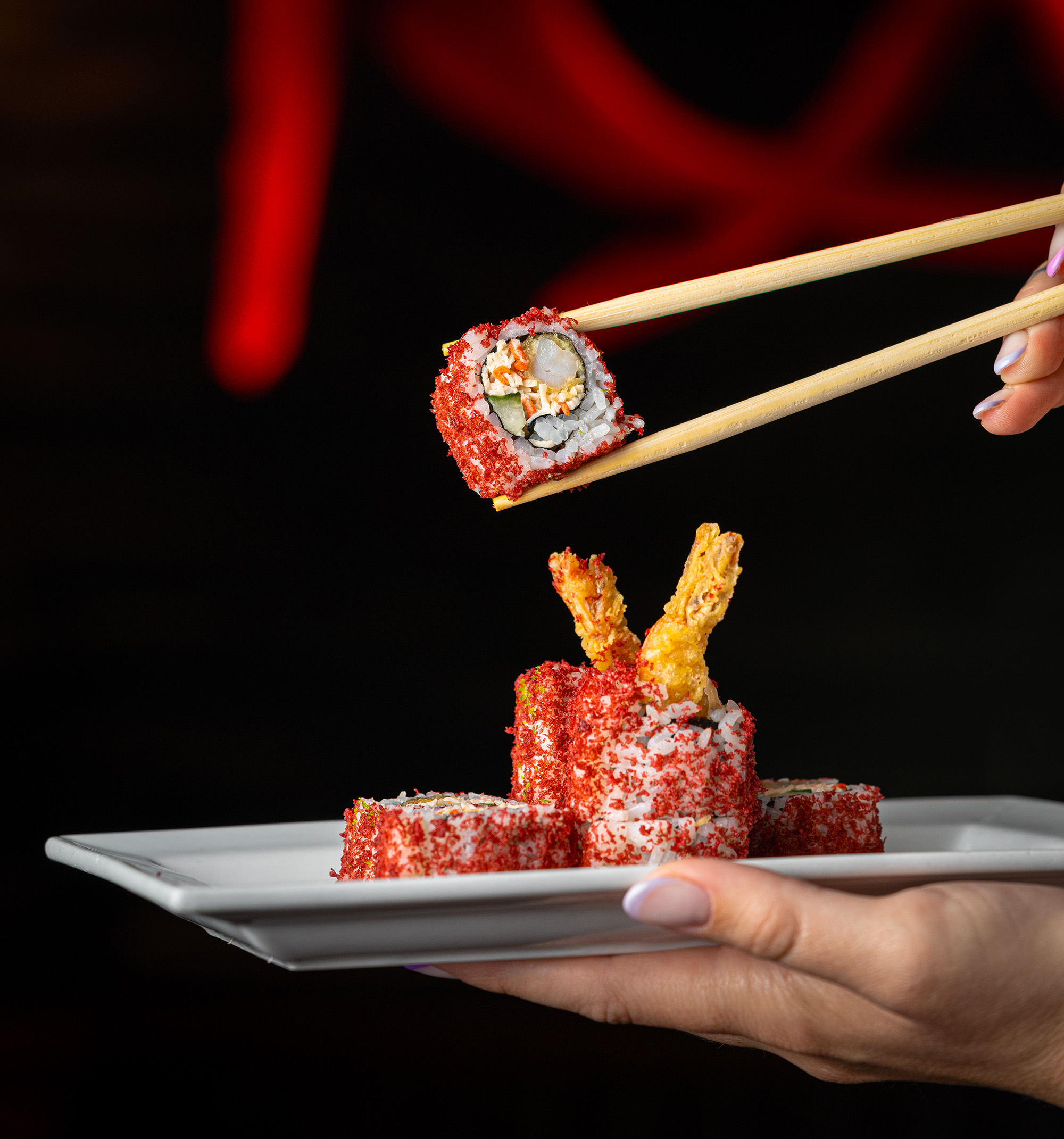 Sushi rolls coated in red crumbs on a white plate, with one piece held above by chopsticks.