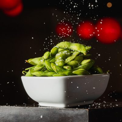 A white bowl filled with edamame is sprinkled with salt, set against a dark background with red lights.