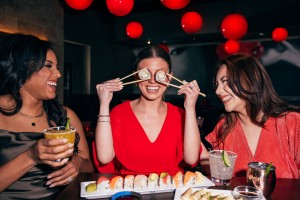 Three women at a table enjoying sushi, cocktails, and drinks in a restaurant with red pendant lights.