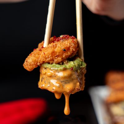 A sushi roll topped with fried shrimp, guacamole, and creamy sauce held by chopsticks.