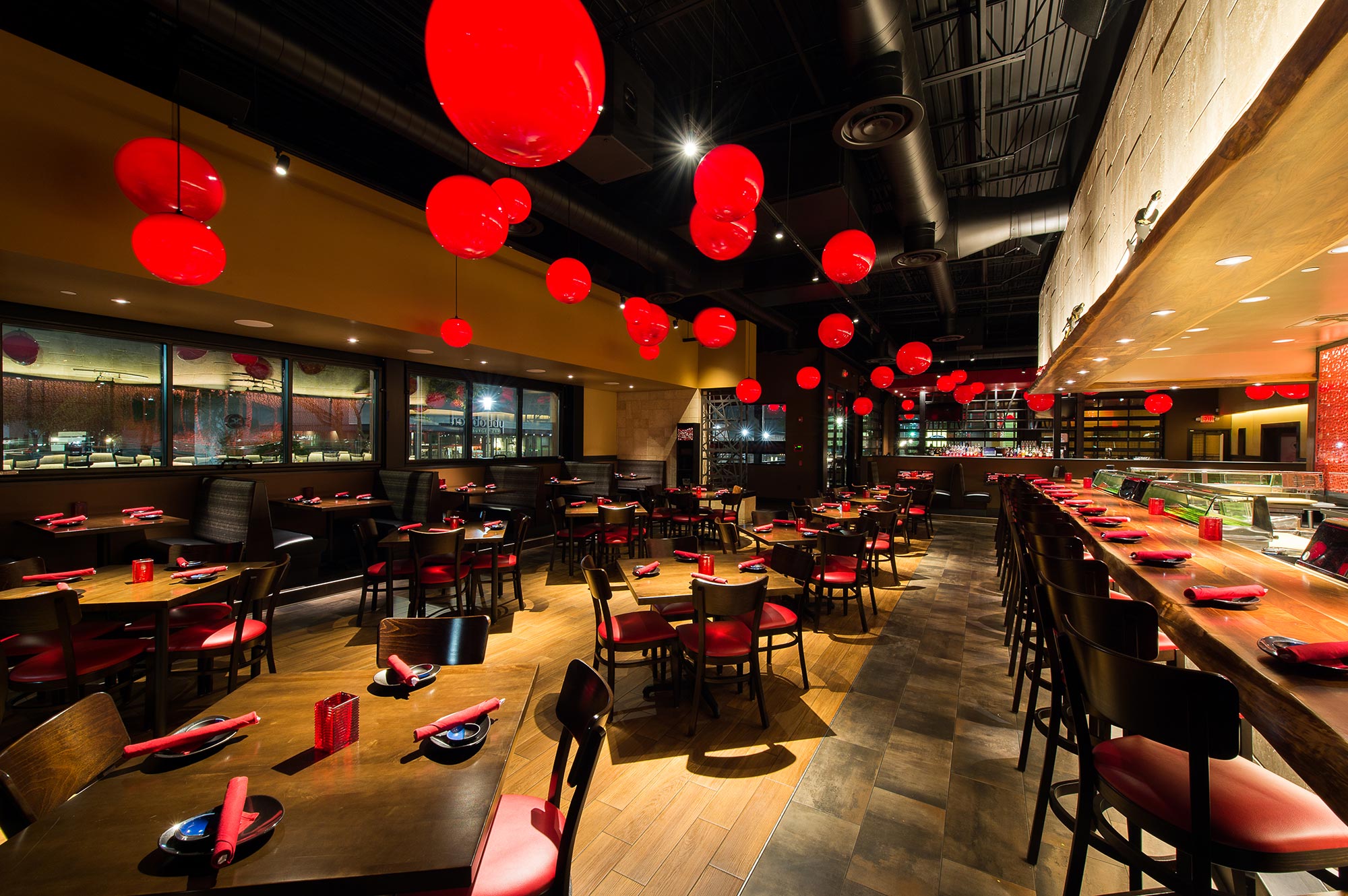 A restaurant interior features red lantern lights, wooden tables with red napkins, and a long sushi bar with counter seating.