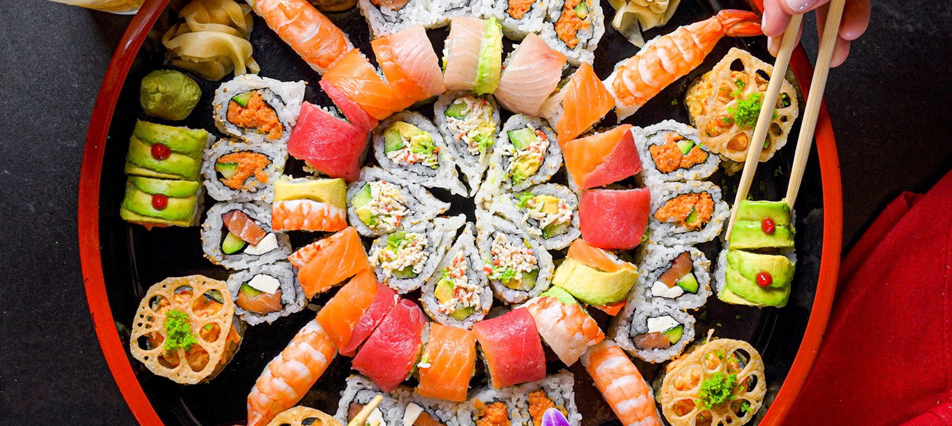 An assorted sushi platter features sushi rolls, nigiri, wasabi, pickled ginger, and lotus chips, with chopsticks picking up a piece.